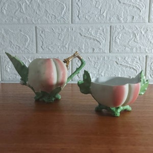 May include: A vintage ceramic creamer and sugar bowl set. The creamer has a handle with gold accents and a spout. Both pieces are white with pink and green accents, resembling a peach. The sugar bowl has a matching design.