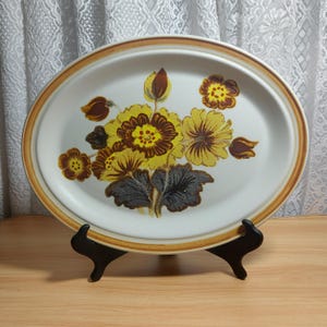 May include: Oval serving platter with a floral design. The white ceramic platter features a brown and yellow floral pattern in the centre, with a brown border. The platter is displayed on a black stand.