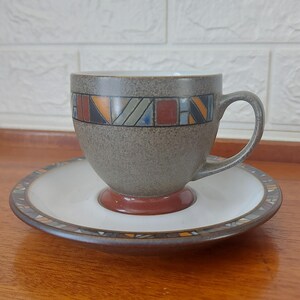 May include: A ceramic teacup and saucer set. The cup is grey with a geometric pattern in orange, blue, and red. The saucer is white with a matching patterned border. The cup sits on a brown base.