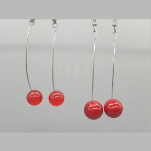 May include: Two pairs of drop earrings featuring long, thin, silver-toned metal wires that curve slightly. Each wire terminates in a single, smooth, glossy, spherical red bead. The earrings are suspended from standard silver-toned metal earring hooks.