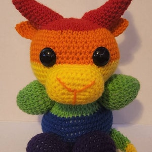 May include: A crocheted rainbow colored plush toy with horns, a tail, and large black eyes. The toy is sitting on a white surface.