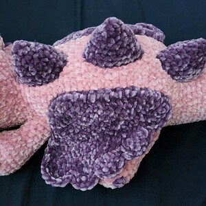 May include: A plush pink and purple dragon-shaped pillow. The dragon has a soft, textured body with purple accents on its back, wings, and head. The dragon has black button eyes and a tail.