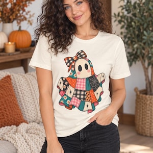 Può includere: Una t-shirt a maniche corte color crema con una grafica centrale raffigurante un fantasma composto da vari motivi patchwork colorati, tra cui quadri, pois e disegni floreali, completato da un fiocco a quadri abbinato sulla testa.