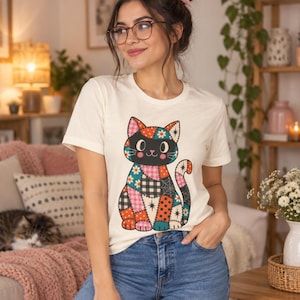 Puede incluir: Una camiseta de manga corta color crema con un gráfico de un gato sentado compuesto por varios patrones de patchwork coloridos, incluyendo lunares, cuadros y diseños florales, combinada con vaqueros azules y un accesorio para el pelo rosa.