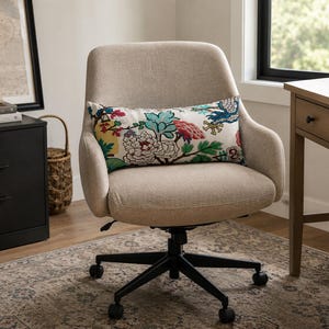 May include: A beige upholstered office chair with a black five-point base on casters. A rectangular lumbar pillow rests on the seat, featuring a white background with a colorful floral pattern in shades of teal, pink, yellow, and green.