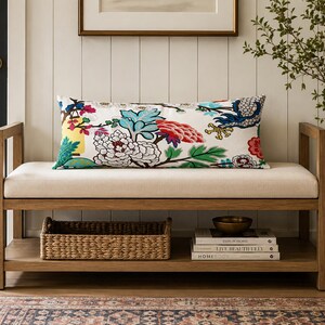 May include: A rectangular lumbar pillow featuring a white background with a vibrant, multi-colored floral and bird pattern in shades of red, blue, yellow, and green. The pillow is positioned on a light-colored upholstered wooden bench with a lower shelf.