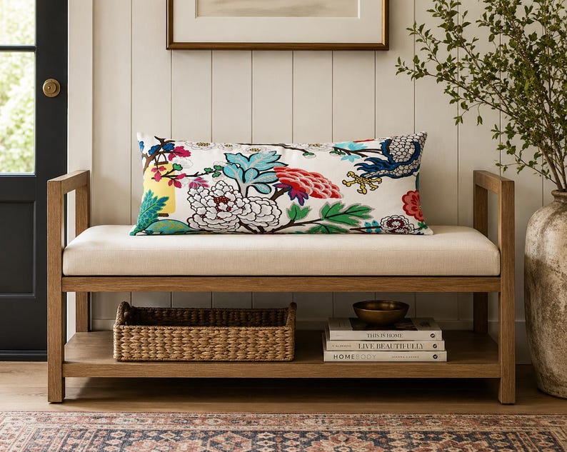May include: A rectangular lumbar pillow featuring a white background with a vibrant, multi-colored floral and bird pattern in shades of red, blue, yellow, and green. The pillow is positioned on a light-colored upholstered wooden bench with a lower shelf.