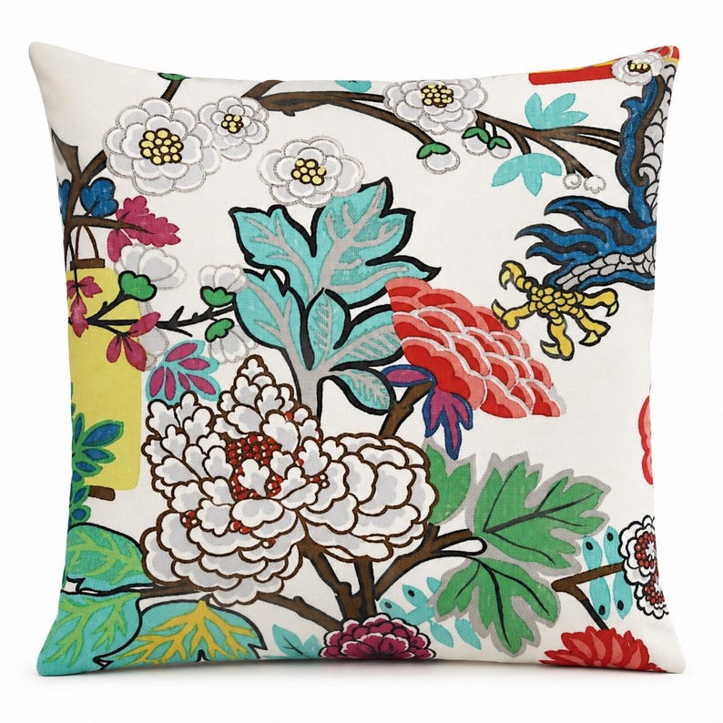 May include: A square throw pillow featuring a white background with a vibrant, multi-colored botanical print. The design includes large white, red, and purple flowers, green leaves, and blue branches, accented by a yellow rectangular shape.