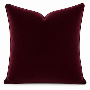 May include: A square throw pillow in a deep burgundy velvet fabric. The pillow features a smooth, solid-colored surface with a distinct piped edge detail along the perimeter, creating a clean, structured silhouette against a plain white background.