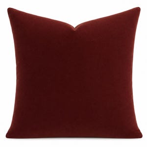 May include: A square throw pillow in a deep, solid burgundy red color. The fabric has a soft, matte texture with a slight nap. The pillow features a distinct karate-chop style indentation at the top center, creating a structured, pointed shape.