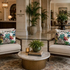 May include: Two cream-colored armchairs with dark brown wooden spindle frames and floral patterned throw pillows featuring teal, red, and yellow accents. A round marble-topped coffee table with a gold fluted base sits between them.