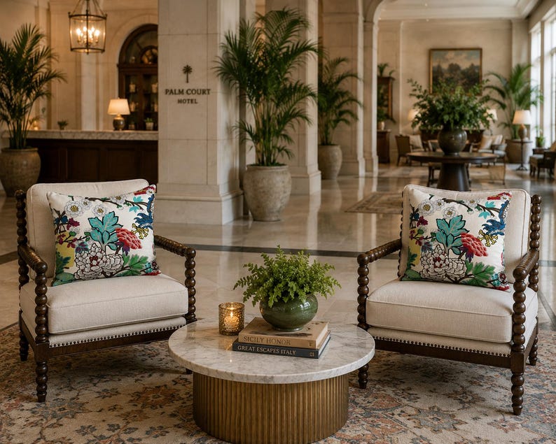 May include: Two cream-colored armchairs with dark brown wooden spindle frames and floral patterned throw pillows featuring teal, red, and yellow accents. A round marble-topped coffee table with a gold fluted base sits between them.