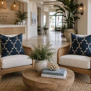 May include: Two armchairs with light-coloured cushions and dark blue patterned throw pillows, featuring woven cane sides and wooden frames. A round wooden coffee table sits between them, holding a potted green plant and two stacked books.