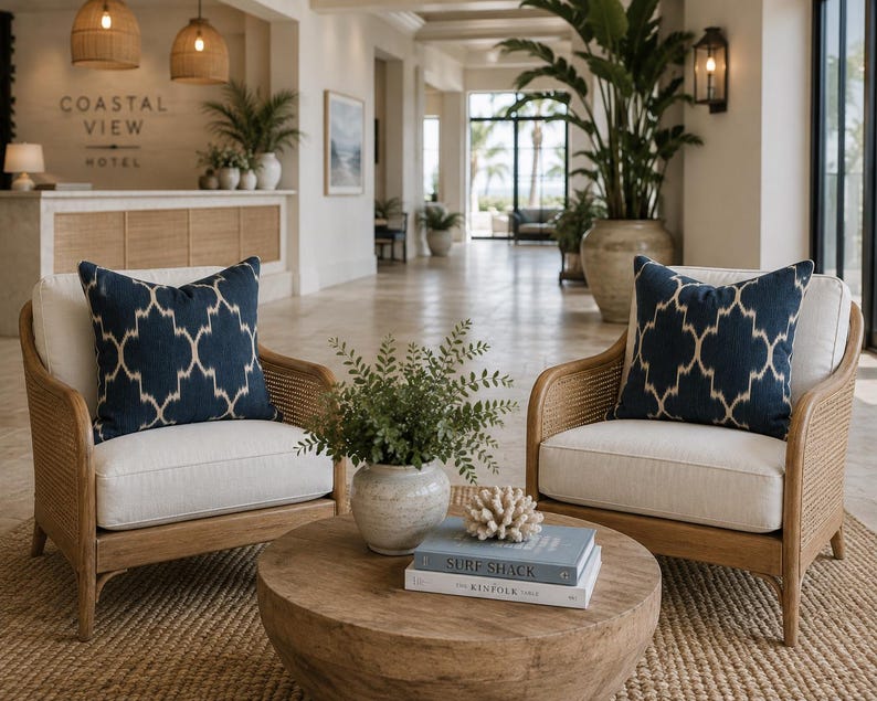 May include: Two armchairs with light-coloured cushions and dark blue patterned throw pillows, featuring woven cane sides and wooden frames. A round wooden coffee table sits between them, holding a potted green plant and two stacked books.