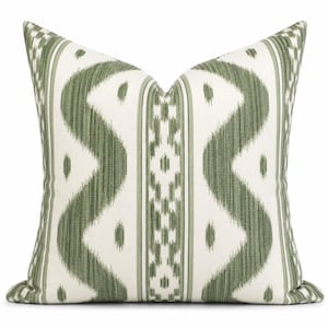 May include: A square throw pillow featuring a white base with a repeating olive green ikat-style pattern. The design includes vertical wavy lines and a central geometric column with small diamond shapes, creating a symmetrical, textured appearance.