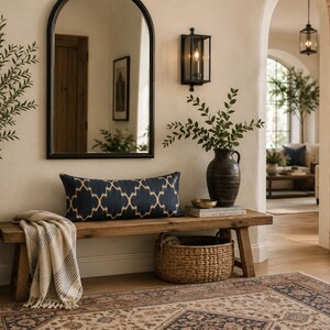 May include: A rectangular navy blue lumbar cushion with a cream-coloured geometric lattice pattern sits on a rustic wooden bench. The bench is positioned in an entryway featuring a large arched mirror, a dark vase with greenery, and a woven basket.