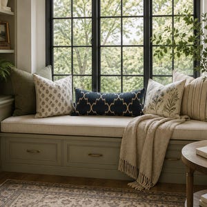 May include: A built-in window seat with sage green drawers and a beige cushion, featuring a collection of decorative pillows in olive green, cream with floral patterns, and a central navy blue lumbar pillow with a white geometric lattice design.