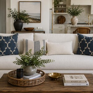 May include: A cream-coloured sofa featuring two navy blue patterned throw pillows, one striped lumbar pillow, and one solid cream lumbar pillow. The sofa is positioned behind a wooden coffee table holding a round woven tray with a plant.