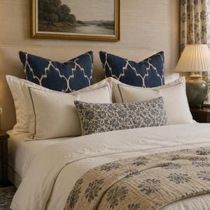May include: A bed featuring a cream-coloured headboard, two large navy blue pillows with a white geometric pattern, two cream pillows with thin dark borders, a long rectangular pillow with a blue floral print, and a cream quilt with blue floral motifs.