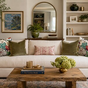 May include: A cream-colored sofa featuring two floral patterned throw pillows, two solid olive green velvet pillows, and one rectangular patterned lumbar pillow. A rustic wooden coffee table sits in front with a book titled The Garden Book.