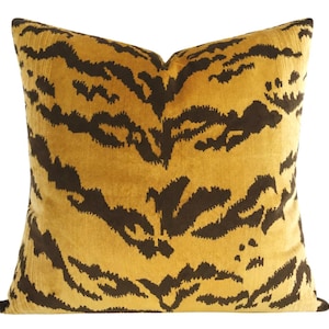 May include: A square throw pillow featuring a mustard yellow base color with a dark brown, irregular, horizontal tiger-stripe pattern. The fabric has a soft, velvet-like texture, and the design covers the entire front surface of the pillow.