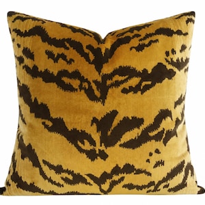 May include: A square throw pillow featuring a mustard yellow base color with a dark brown, abstract tiger stripe pattern. The fabric has a soft, velvet-like texture with a visible nap that creates subtle variations in the depth of the colors.
