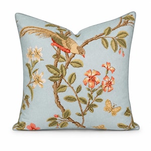 May include: A square throw pillow featuring a light blue background with a printed pattern of a long-tailed bird perched on a brown branch, surrounded by orange and white flowers, green leaves, and small yellow butterflies, finished with light blue piping.