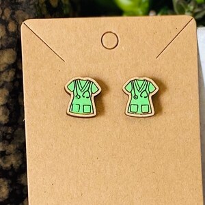 May include: A pair of wooden earrings shaped like a green medical scrub top with a stethoscope. The earrings are on a brown card.