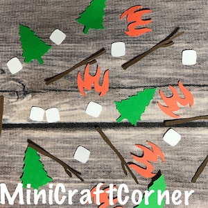 May include: A collection of paper cutouts arranged on a wood grain surface. The cutouts include green trees, orange flames, brown sticks, and white marshmallows. The text "MiniCraftCorner" is at the bottom. The theme is camping and outdoor activities.
