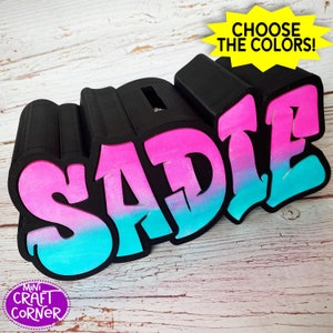 May include: A 3D printed name sign with the name "SADIE" in a graffiti style. The letters are black with a pink and blue ombre effect. The sign is on a wooden surface.