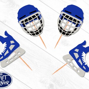 May include: Blue hockey helmet and ice skate cupcake toppers. The helmet has a white and black cage, and the skate has white laces and a gray blade. The toppers are on wooden sticks. The Mini Craft Corner logo is in the lower left corner.