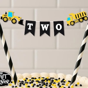 May include: A cake topper with a construction theme. The topper features a banner that spells "TWO" with a yellow and black dump truck and a yellow and black excavator. The topper is supported by two black and white striped straws.