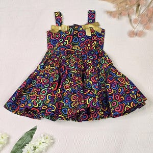 May include: A colorful dress with a vibrant pattern of multi-colored shapes on a black background. The dress features gold bows on the straps and a flared skirt. The dress is designed for a child.