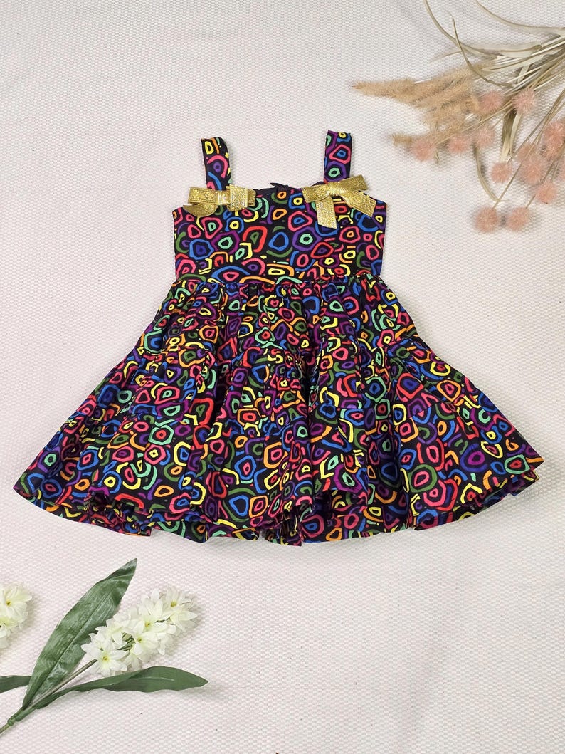 May include: A colorful dress with a vibrant pattern of multi-colored shapes on a black background. The dress features gold bows on the straps and a flared skirt. The dress is designed for a child.