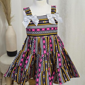 May include: A sleeveless dress with a vibrant pattern of pink, yellow, black, and white geometric shapes. The dress features silver bows on the straps and a tiered skirt. The dress is displayed on a mannequin.