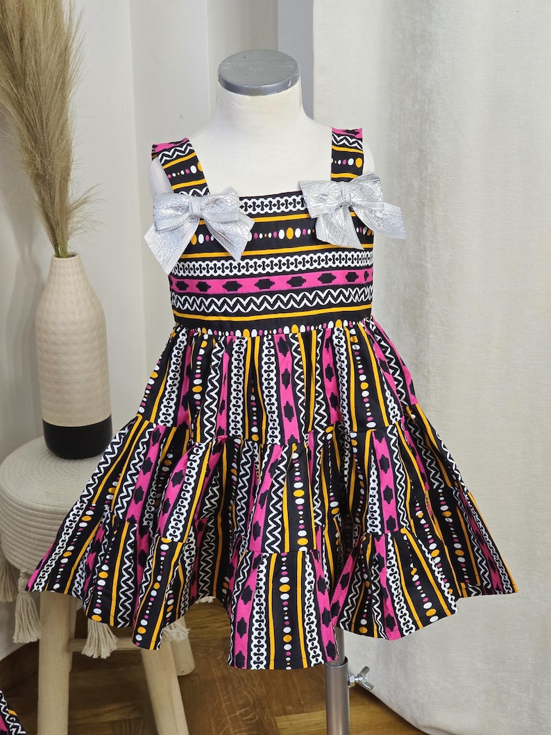 May include: A sleeveless dress with a vibrant pattern of pink, yellow, black, and white geometric shapes. The dress features silver bows on the straps and a tiered skirt. The dress is displayed on a mannequin.