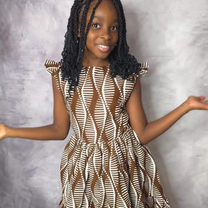 Könnte beinhalten: Braunes und weiß gemustertes Kleid mit Rüschensaum. Das Kleid hat einen runden Ausschnitt und Flügelärmel. Das Kleid wird von einer Person mit geflochtenem Haar getragen.