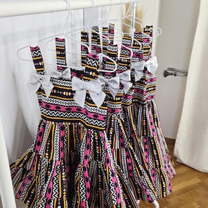 May include: Several dresses with a vibrant, patterned design hang on a white rack. The dresses feature a black, pink, yellow, and white geometric print. Each dress has a silver bow at the neckline and a tiered skirt.