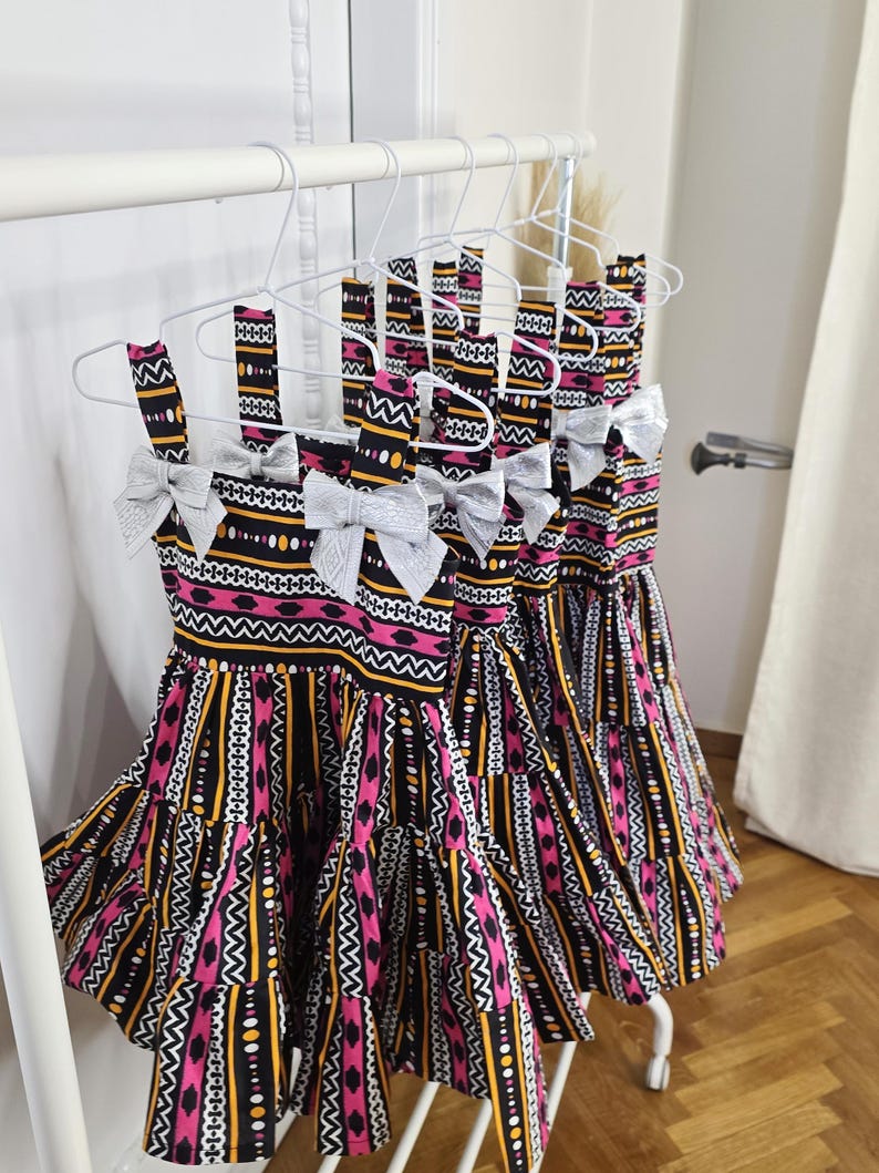 May include: Several dresses with a vibrant, patterned design hang on a white rack. The dresses feature a black, pink, yellow, and white geometric print. Each dress has a silver bow at the neckline and a tiered skirt.