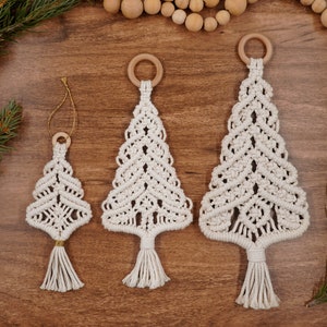 May include: Three white macrame Christmas tree ornaments with wooden rings at the top and tassels at the bottom. The ornaments are hanging on a wooden surface.