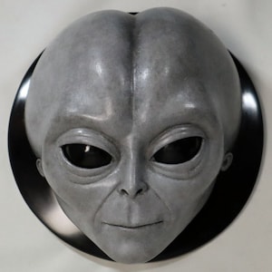 May include: A gray alien head sculpture with large black eyes and a slightly smiling expression. The sculpture is mounted on a black circular base.