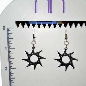 Peut inclure: Une paire de boucles d'oreilles noires en forme d'éclat de soleil exposée sur un présentoir blanc. Les boucles d'oreilles sont en métal foncé et présentent un motif circulaire avec des pointes rayonnantes. Le présentoir a une règle imprimée sur le côté pour la mesure.