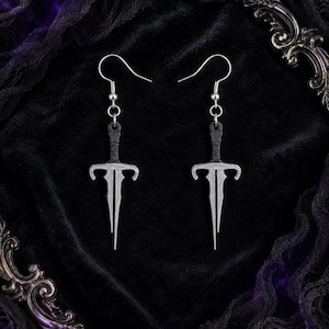 May include: A pair of silver-toned earrings shaped like daggers with black wrapped handles. Each earring features a long, pointed blade, a crossguard, and a hook-style ear wire for attachment. The design is symmetrical and metallic in appearance.