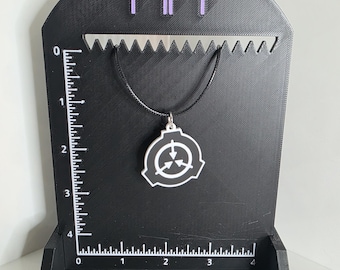 Op de afbeelding: Een zwart-witte SCP Foundation logo hanger aan een zwart koord, gepresenteerd op een zwarte 3D-geprinte standaard met een liniaal tot 10 cm. De standaard heeft een paars 'nh' logo aan de bovenkant en een gekartelde rand voor sieraden.