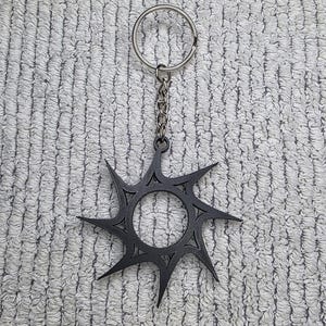 May include: A dark gray sunburst-shaped keychain pendant with a silver chain and keyring. The pendant has a circular center with pointed rays extending outwards. The keychain is displayed on a textured, light gray surface.