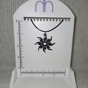 May include: A black sunburst pendant necklace on a black cord. The pendant is displayed on a white stand with a ruler on the side. The stand has the letters "nh" in purple at the top.