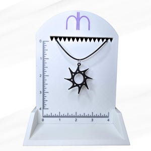 Peut inclure: Un collier pendentif noir en forme d'éclat de soleil présenté sur un présentoir blanc avec une règle. Le collier a un cordon noir et un pendentif en forme de soleil avec des rayons pointus. Le présentoir porte les lettres "nh" en violet.