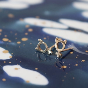 May include: Two silver earrings in the shape of the female gender symbol on a blue and white background.