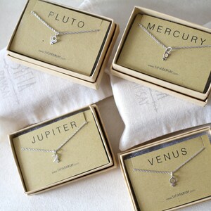 May include: Four small boxes with brown lids and white interiors, each containing a silver necklace with a charm representing a planet. The boxes are labeled "Pluto", "Mercury", "Jupiter", and "Venus".