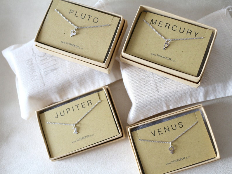 May include: Four small boxes with brown lids and white interiors, each containing a silver necklace with a charm representing a planet. The boxes are labeled "Pluto", "Mercury", "Jupiter", and "Venus".